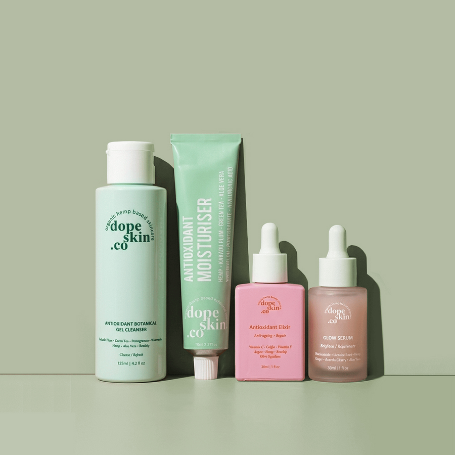 Four skincare products from dote skin co. arranged on a muted sage green background: a light green bottle of gel cleanser, a mint green tube of moisturiser, a pink bottle of body wash, and a pink dropper bottle of serum.